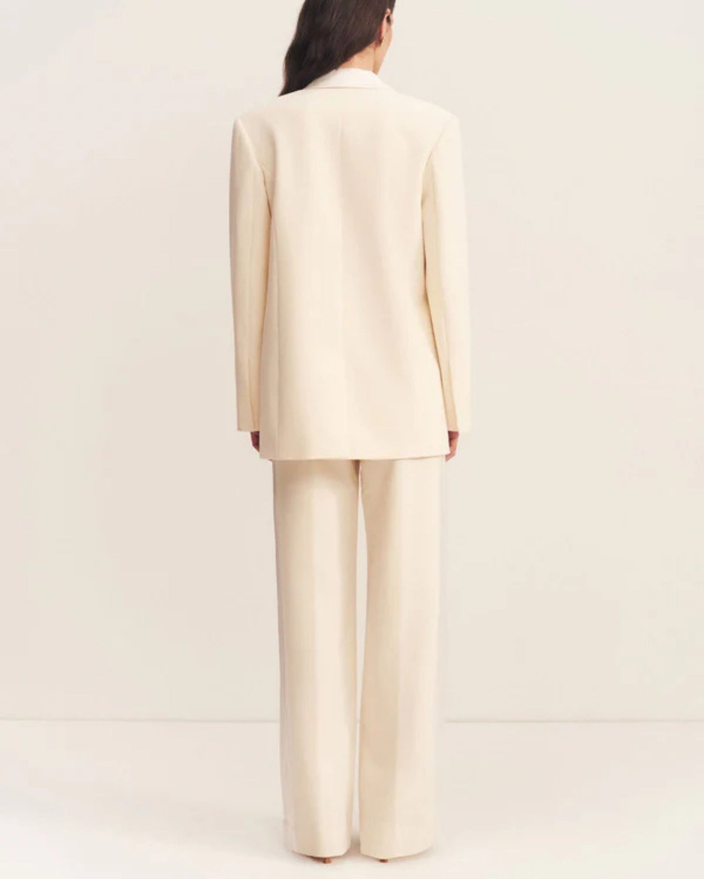 Back view, a double-breasted blazer in soft rice with contrast satin lapel, collar, and flap pockets. Features tailored sleeves, back vent, and structured silhouette. Available at Georgette bridal boutique in Brussels and online at georgettebridal.com.