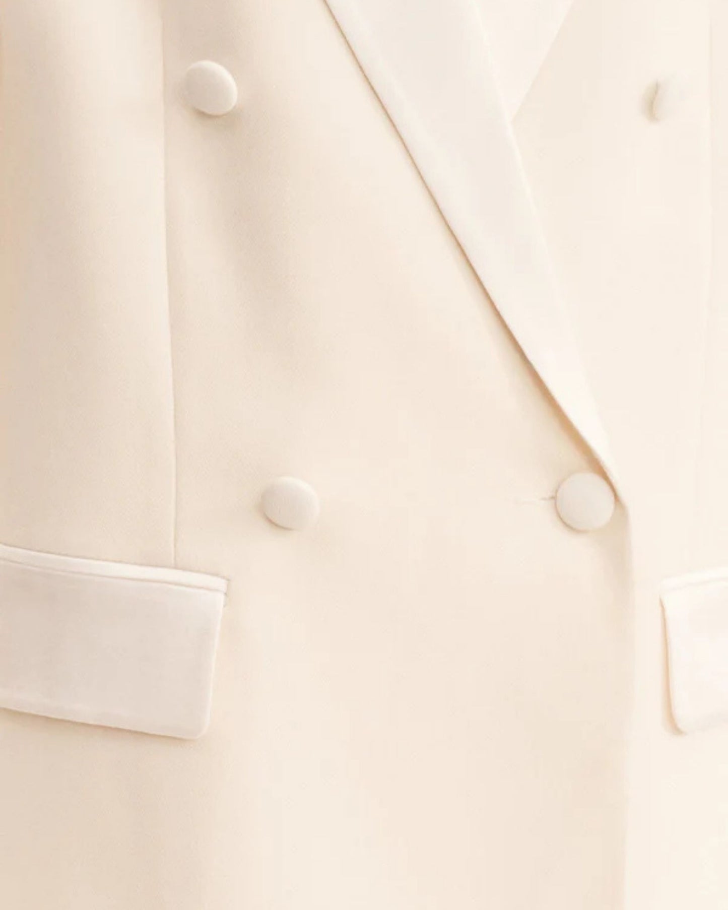 Close-up, double-breasted blazer in soft rice with contrast satin lapel, collar, and flap pockets. Features tailored sleeves, back vent, and structured silhouette. Available at Georgette bridal boutique in Brussels and online at georgettebridal.com.