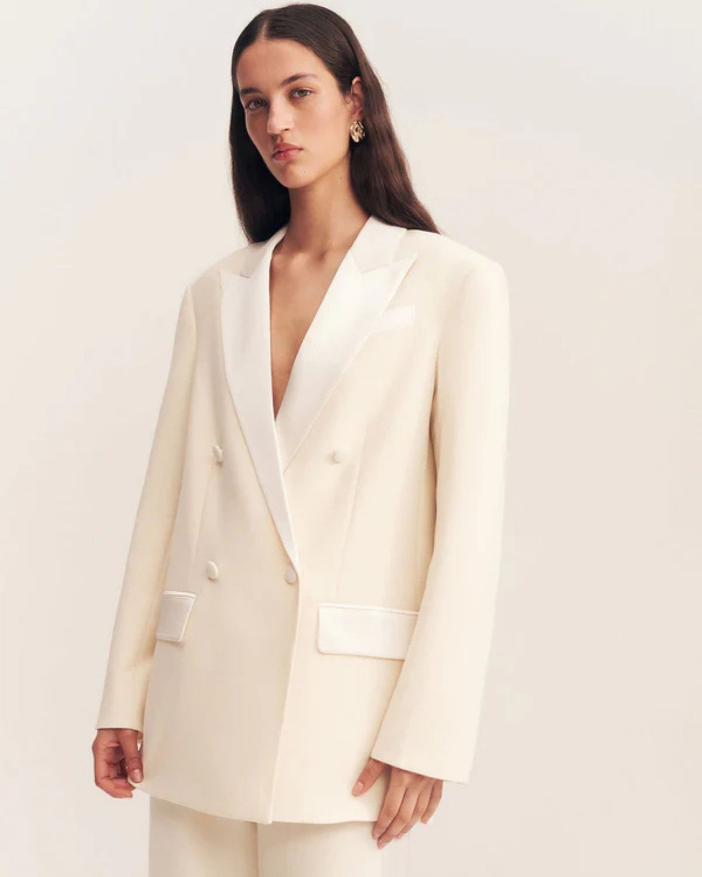 Model wearing a double-breasted blazer in soft rice with contrast satin lapel, collar, and flap pockets. Features tailored sleeves, back vent, and structured silhouette. Available at Georgette bridal boutique in Brussels and online at georgettebridal.com.
