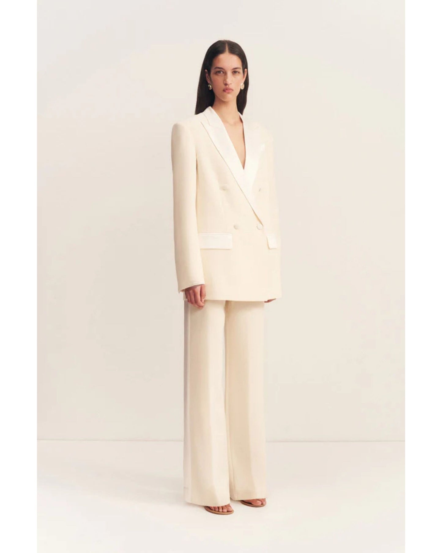 Model wearing a double-breasted blazer in soft rice with contrast satin lapel, collar, and flap pockets. Features tailored sleeves, back vent, and structured silhouette. Available at Georgette bridal boutique in Brussels and online at georgettebridal.com.