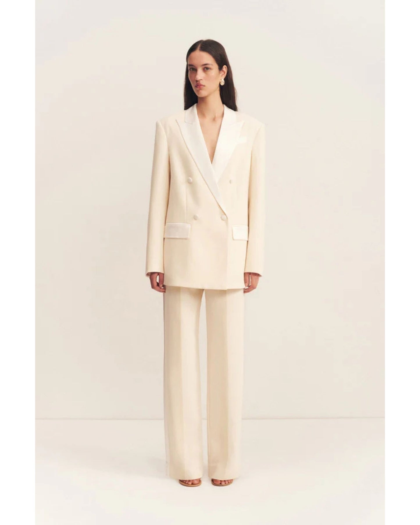 Model wearing a double-breasted blazer in soft rice with contrast satin lapel, collar, and flap pockets. Features tailored sleeves, back vent, and structured silhouette. Available at Georgette bridal boutique in Brussels and online at georgettebridal.com.