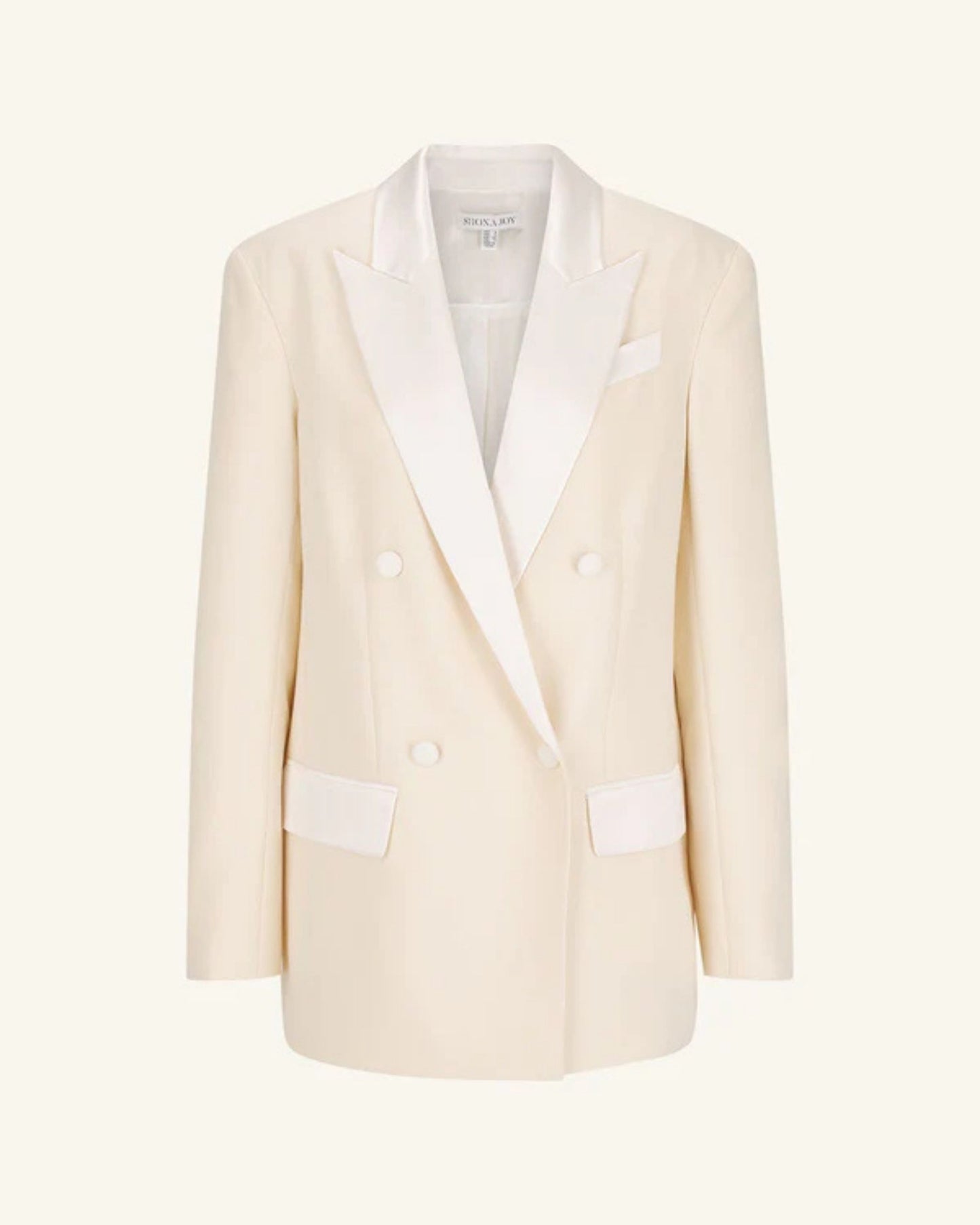 Product shot, a double-breasted blazer in soft rice with contrast satin lapel, collar, and flap pockets. Features tailored sleeves, back vent, and structured silhouette. Available at Georgette bridal boutique in Brussels and online at georgettebridal.com.