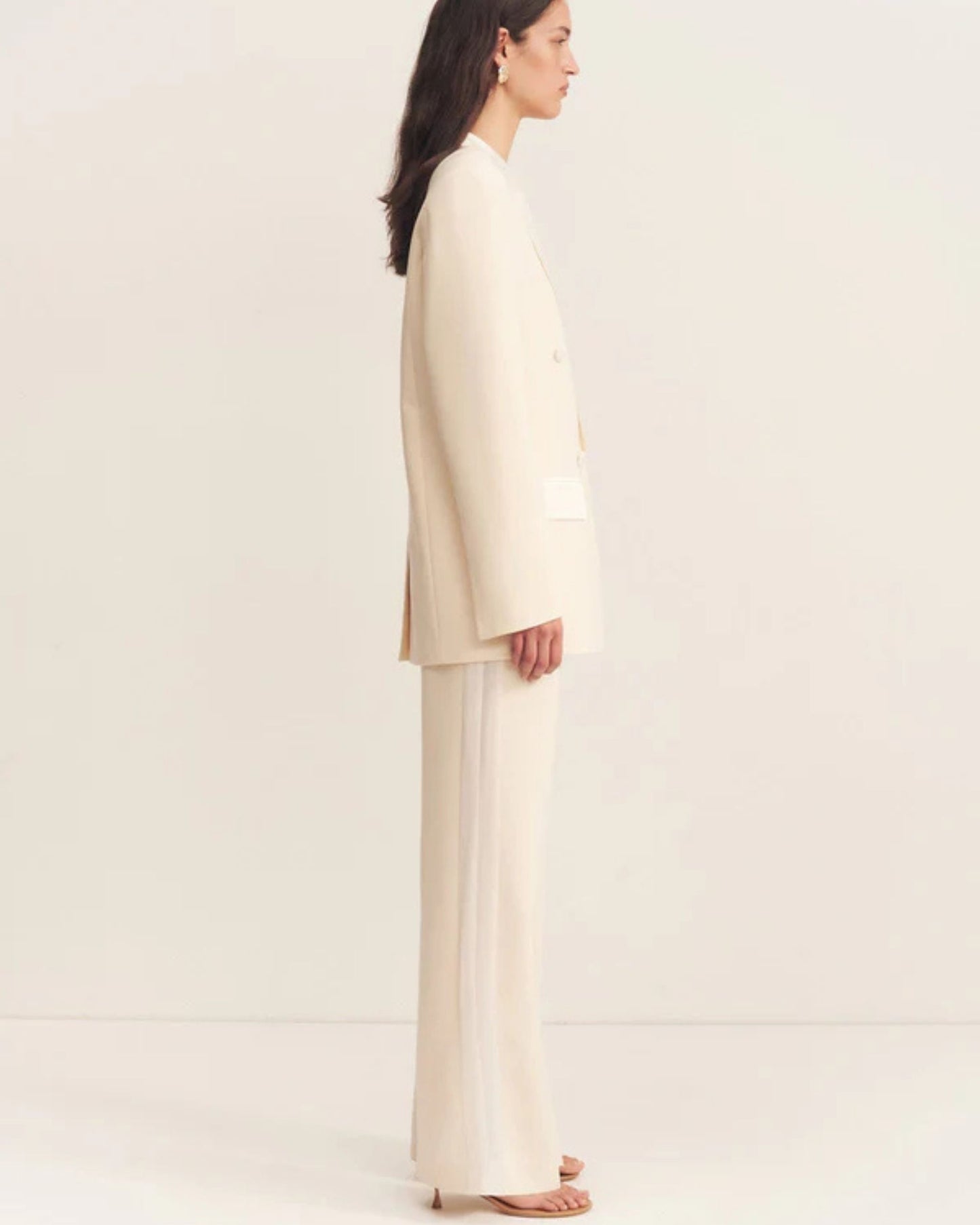 Side view, model wearing a double-breasted blazer in soft rice with contrast satin lapel, collar, and flap pockets. Features tailored sleeves, back vent, and structured silhouette. Available at Georgette bridal boutique in Brussels and online at georgettebridal.com.