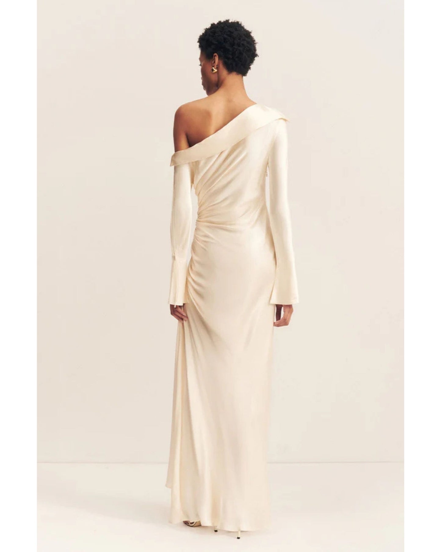 Back view of a cream satin wedding dress with an asymmetrical neckline, ruched side, cuffed sleeves with ties, and a maxi skirt with a front split. Available at Georgette bridal boutique in Brussels and online at georgettebridal.com.