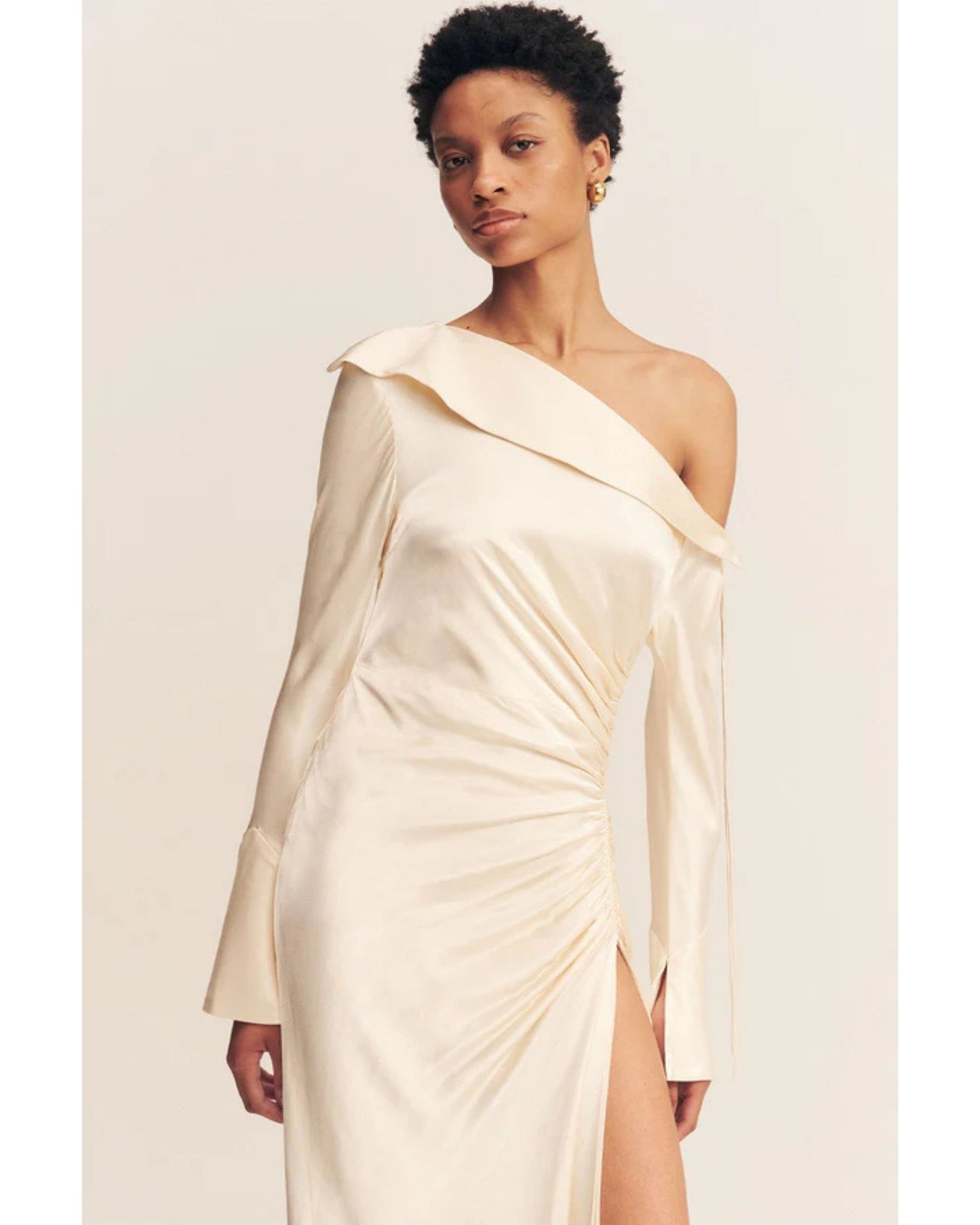 Close-up of a cream satin wedding dress with an asymmetrical neckline, ruched side, cuffed sleeves with ties, and a maxi skirt with a front split. Available at Georgette bridal boutique in Brussels and online at georgettebridal.com.