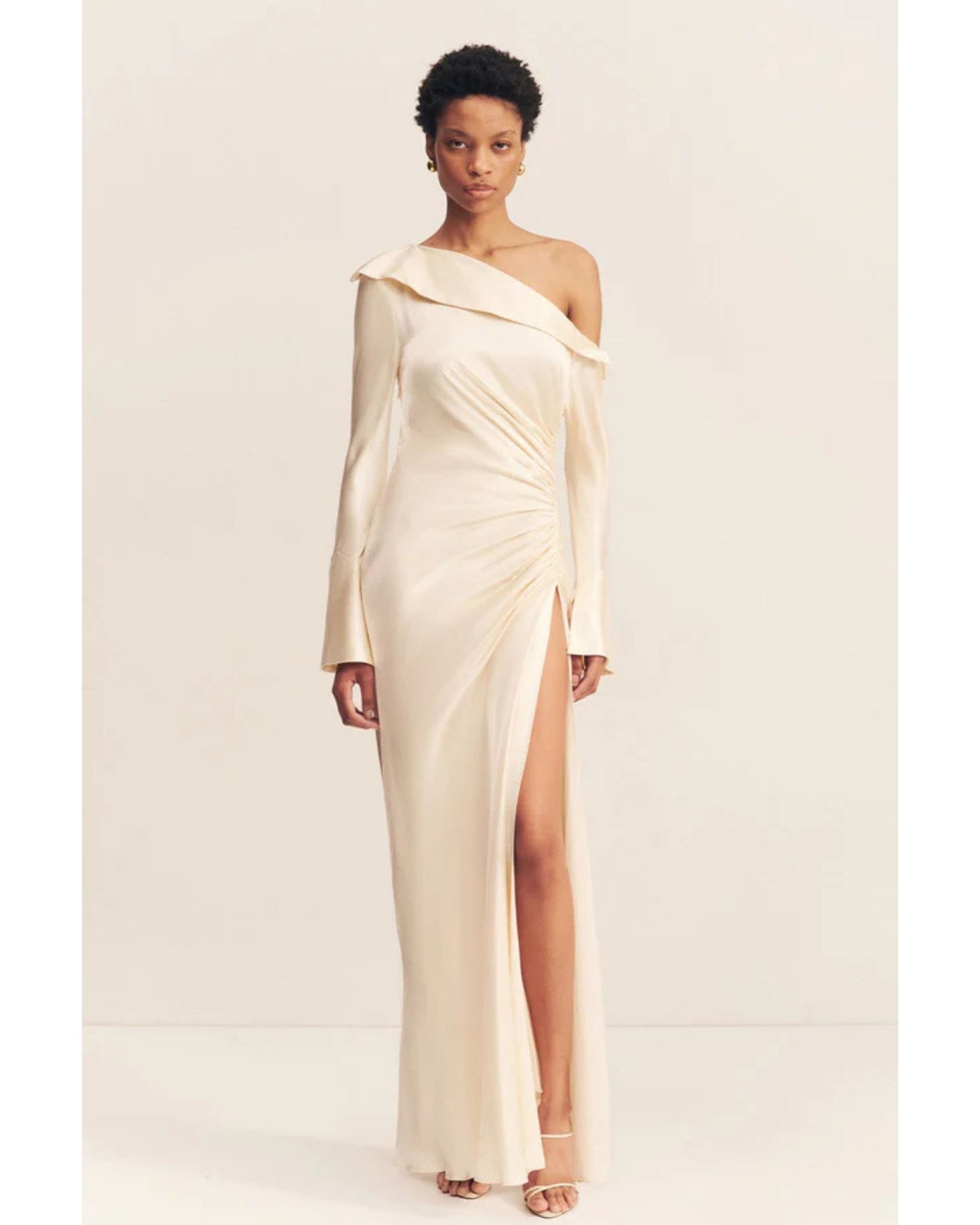 Model wearing a cream satin wedding dress with an asymmetrical neckline, ruched side, cuffed sleeves with ties, and a maxi skirt with a front split. Available at Georgette bridal boutique in Brussels and online at georgettebridal.com