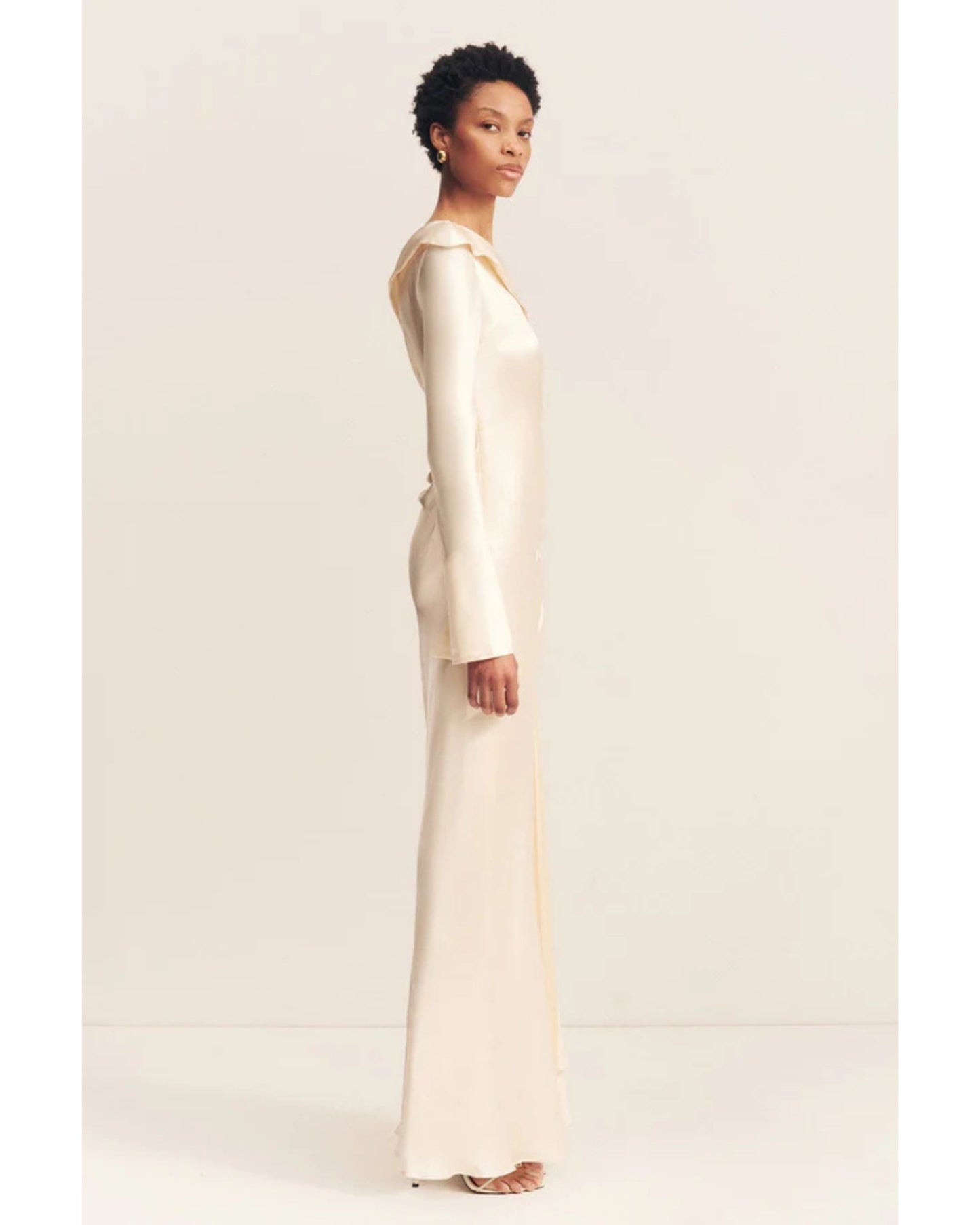Side view of a cream satin wedding dress with an asymmetrical neckline, ruched side, cuffed sleeves with ties, and a maxi skirt with a front split. Available at Georgette bridal boutique in Brussels and online at georgettebridal.com.