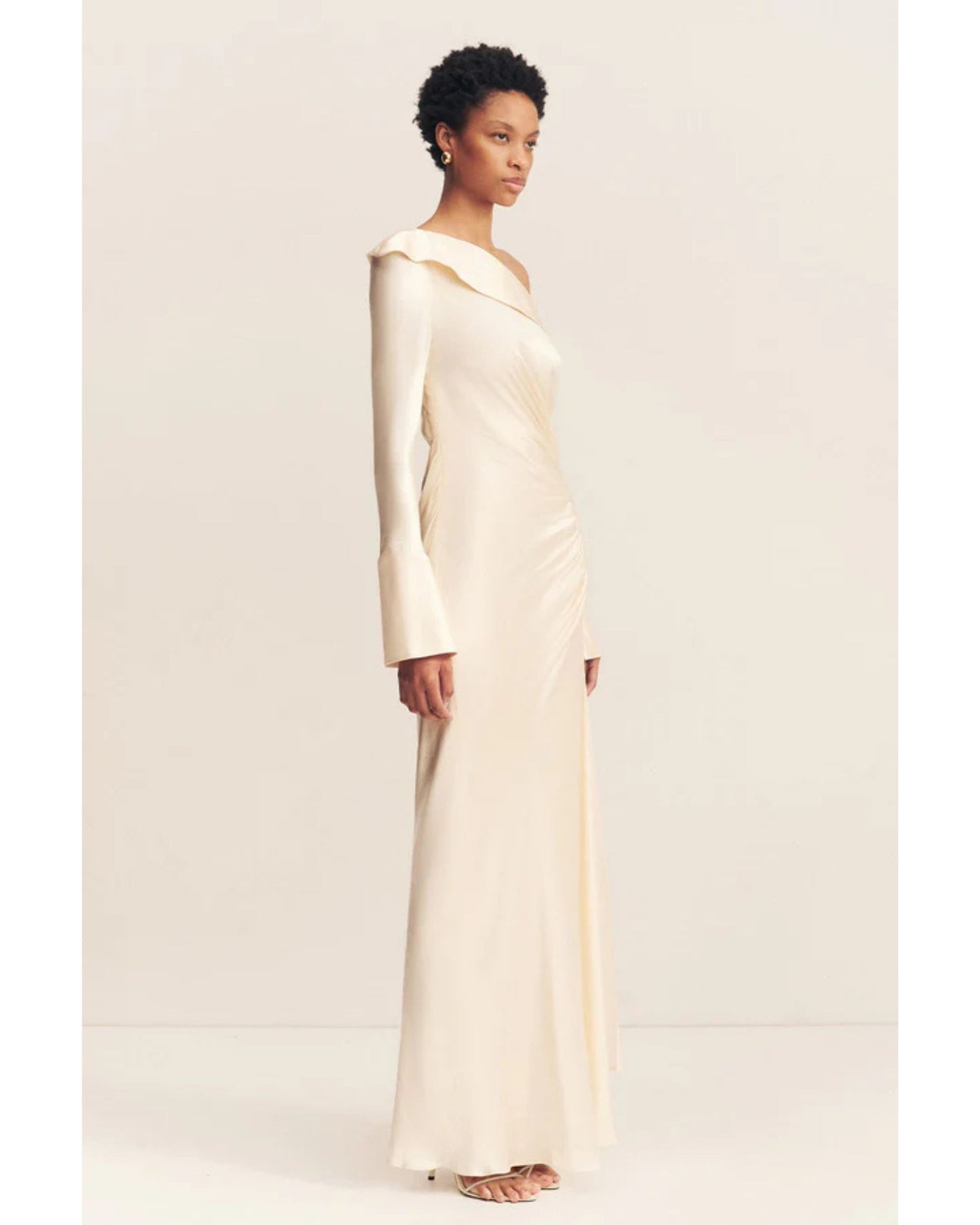 Model wearing a cream satin wedding dress with an asymmetrical neckline, ruched side, cuffed sleeves with ties, and a maxi skirt with a front split. Available at Georgette bridal boutique in Brussels and online at georgettebridal.com.