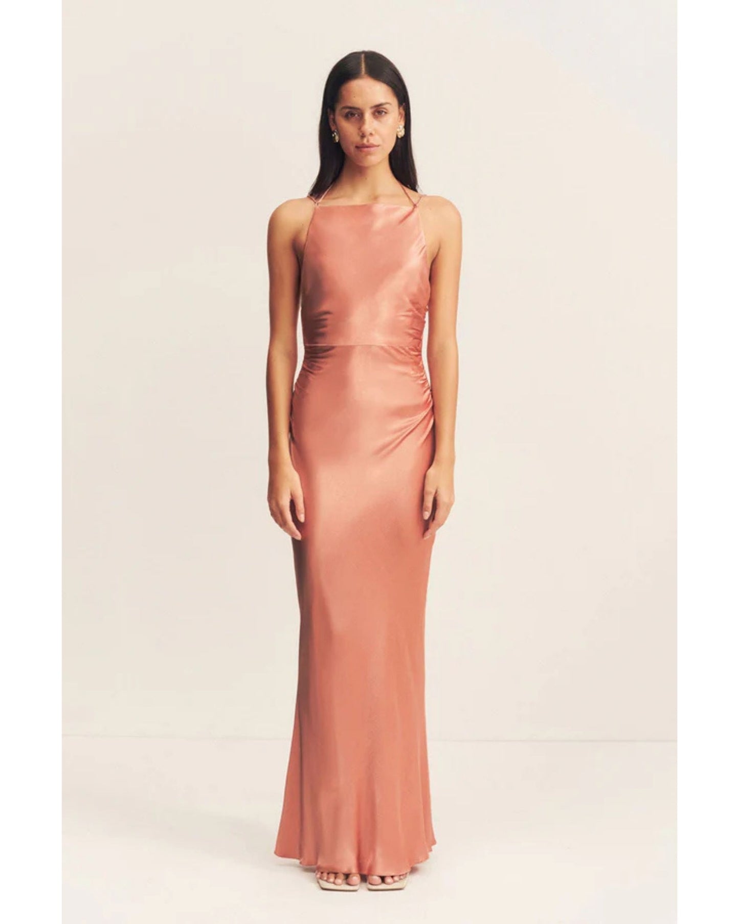 Model wearing an antique rose satin maxi dress with a square high neckline, halter tie, ruched waist, and low open back. Made from sustainable LENZING™ ECOVERO™ viscose. Available at Georgette bridal boutique in Brussels and online at georgettebridal.com.