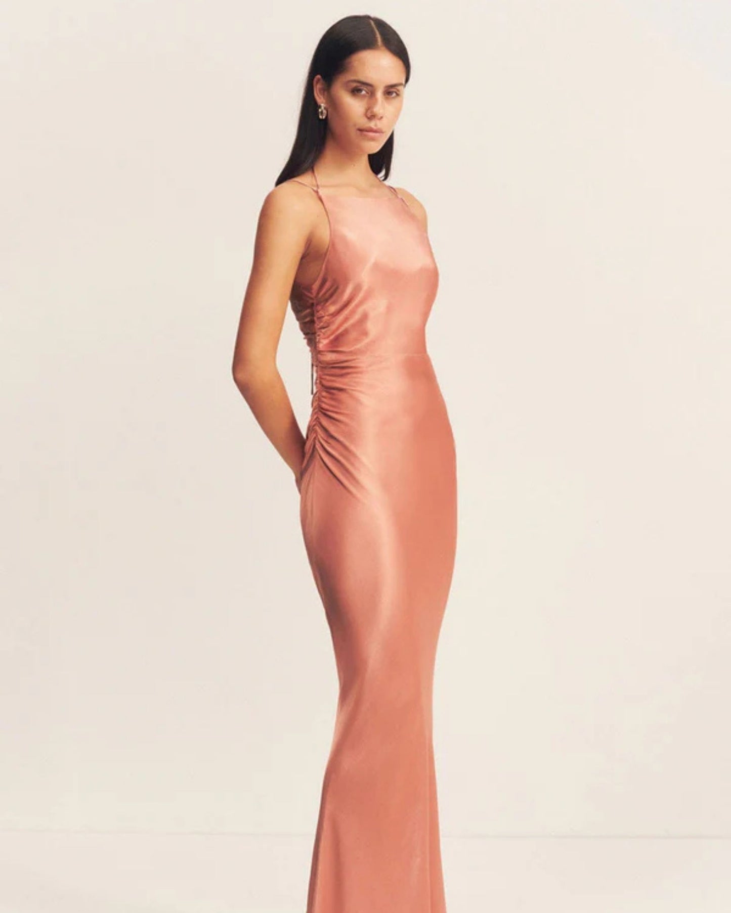 Model wearing an antique rose satin maxi dress with a square high neckline, halter tie, ruched waist, and low open back. Made from sustainable LENZING™ ECOVERO™ viscose. Available at Georgette bridal boutique in Brussels and online at georgettebridal.com.