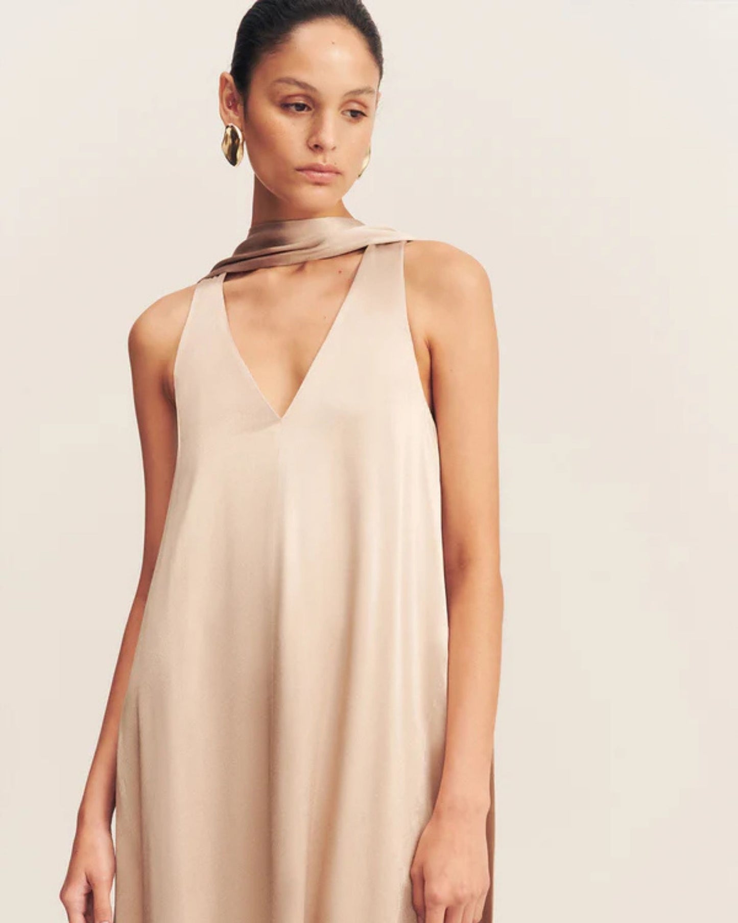 Model wearing an elegant golden maxi dress with a deep plunging neckline and draped scarf detail, made from satin-finish LENZING™ ECOVERO™ viscose for a sophisticated, fluid look. Available at Georgette bridal boutique in Brussels and online at georgettebridal.com.