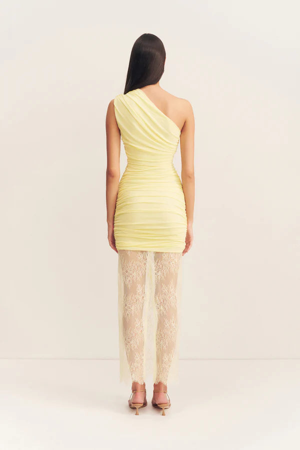 Back view, asymmetrical yellow one-shoulder maxi dress featuring sheer mesh and delicate lace contrast, gathered details, and a flowing lace skirt with a subtle back split. Available at Georgette bridal boutique in Brussels and online at georgettebridal.com.