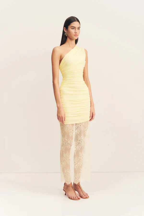 Model wearing an asymmetrical yellow one-shoulder maxi dress featuring sheer mesh and delicate lace contrast, gathered details, and a flowing lace skirt with a subtle back split. Available at Georgette bridal boutique in Brussels and online at georgettebridal.com.