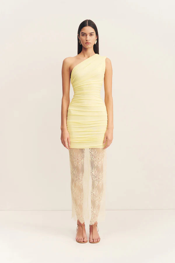 Model wearing an asymmetrical yellow one-shoulder maxi dress featuring sheer mesh and delicate lace contrast, gathered details, and a flowing lace skirt with a subtle back split. Available at Georgette bridal boutique in Brussels and online at georgettebridal.com.