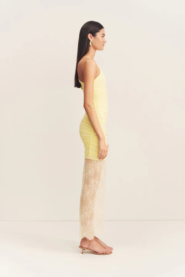 Side view, asymmetrical yellow one-shoulder maxi dress featuring sheer mesh and delicate lace contrast, gathered details, and a flowing lace skirt with a subtle back split. Available at Georgette bridal boutique in Brussels and online at georgettebridal.com.