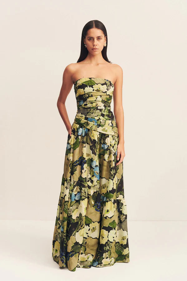 Model wearing a strapless tiered maxi dress in a sheer Botanical Floral print, featuring a softly draped asymmetrical bodice, full tiered skirt, and invisible side zip. Crafted from a linen and TENCEL™ blend. Available at Georgette bridal boutique in Brussels and online at georgettebridal.com.