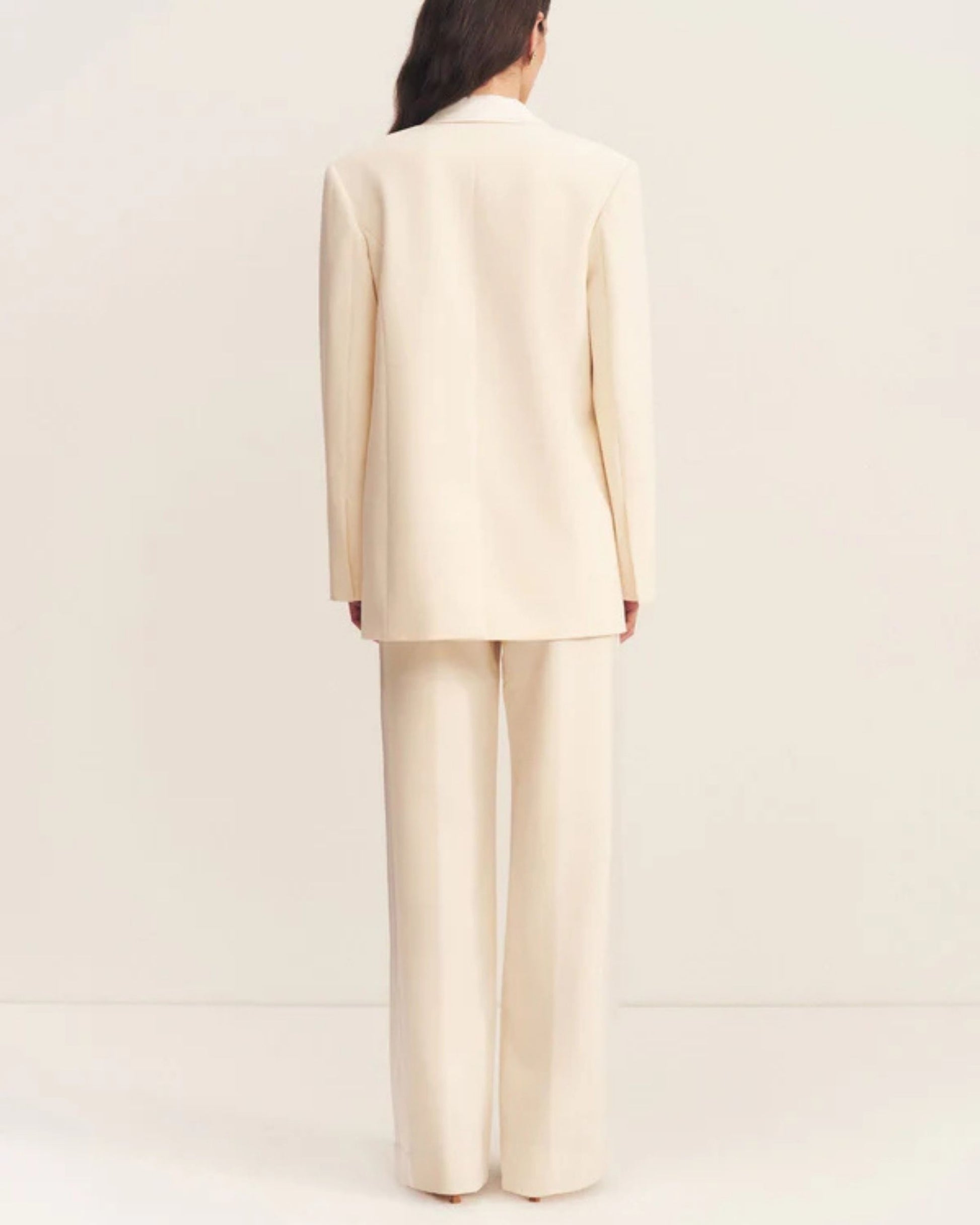 Back view, a double-breasted blazer in soft rice with contrast satin lapel, collar, and flap pockets. Features tailored sleeves, back vent, and structured silhouette. Available at Georgette bridal boutique in Brussels and online at georgettebridal.com.