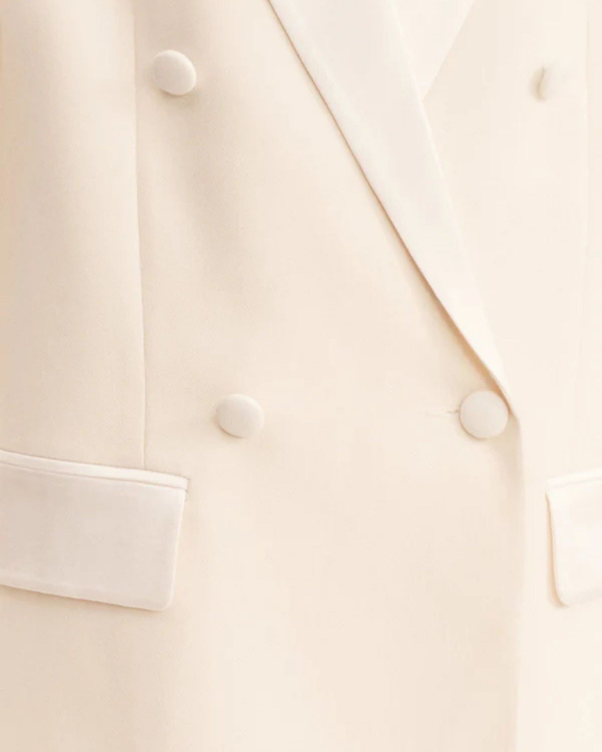 Close-up, double-breasted blazer in soft rice with contrast satin lapel, collar, and flap pockets. Features tailored sleeves, back vent, and structured silhouette. Available at Georgette bridal boutique in Brussels and online at georgettebridal.com.