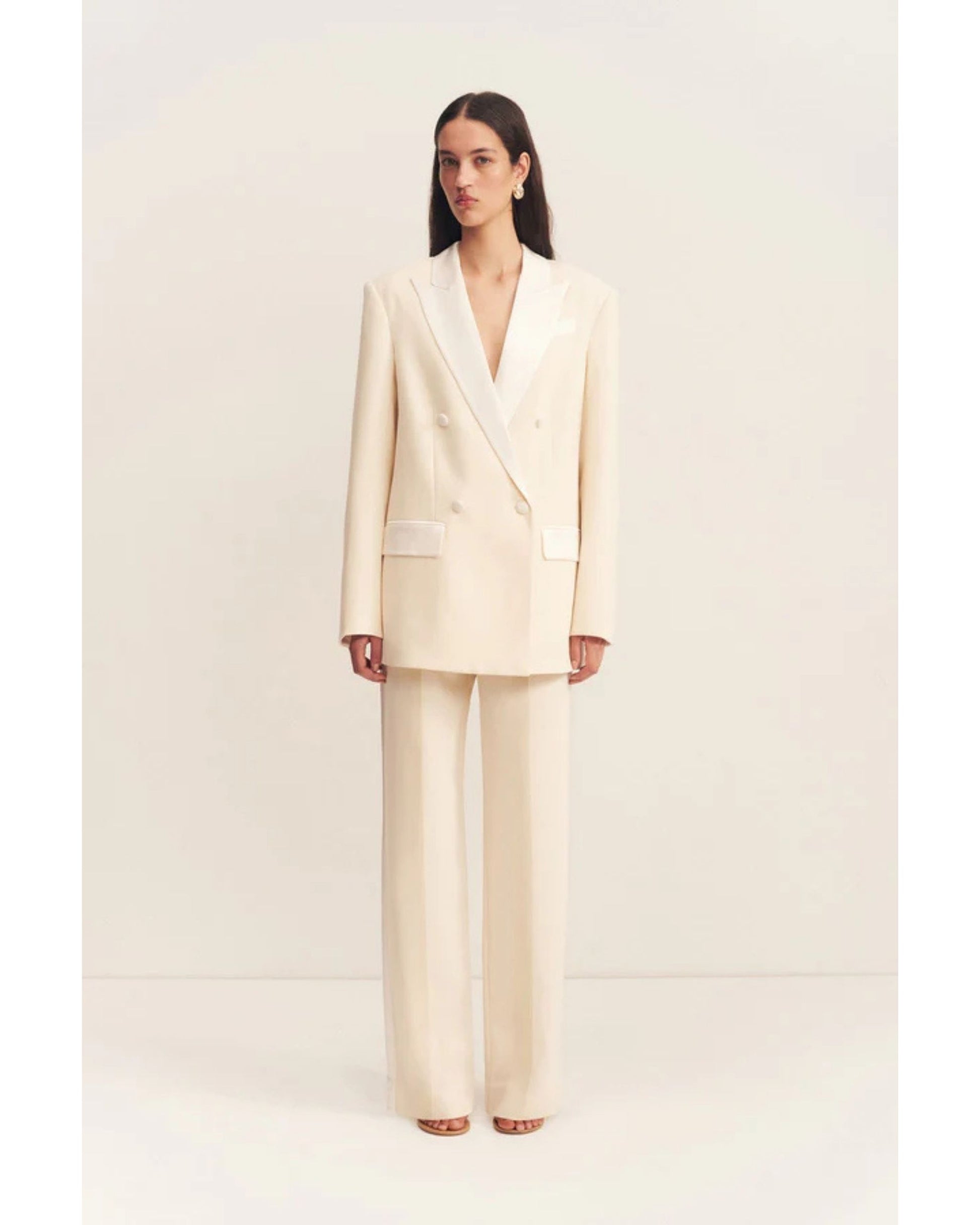 Model wearing a double-breasted blazer in soft rice with contrast satin lapel, collar, and flap pockets. Features tailored sleeves, back vent, and structured silhouette. Available at Georgette bridal boutique in Brussels and online at georgettebridal.com.