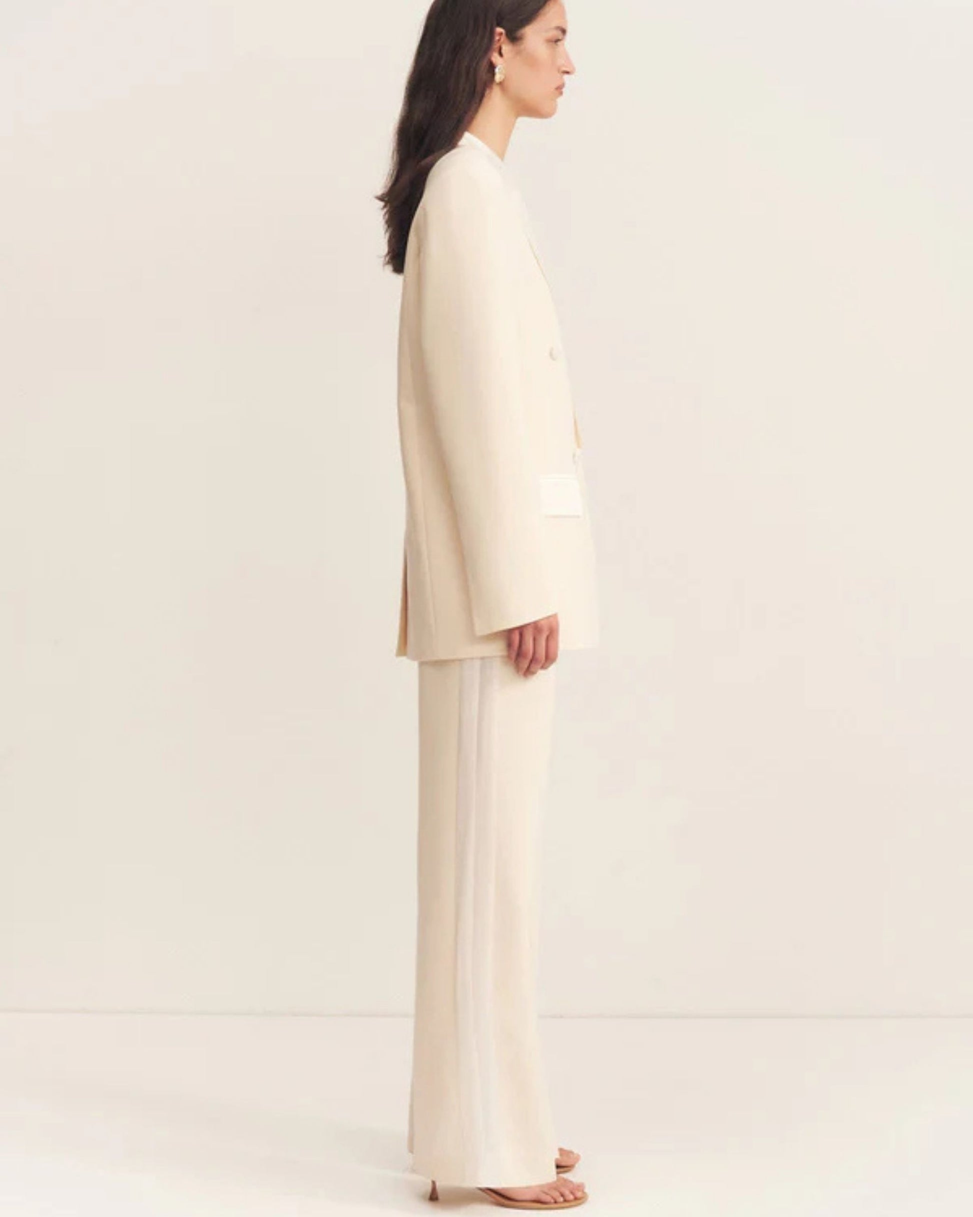Side view, model wearing a double-breasted blazer in soft rice with contrast satin lapel, collar, and flap pockets. Features tailored sleeves, back vent, and structured silhouette. Available at Georgette bridal boutique in Brussels and online at georgettebridal.com.