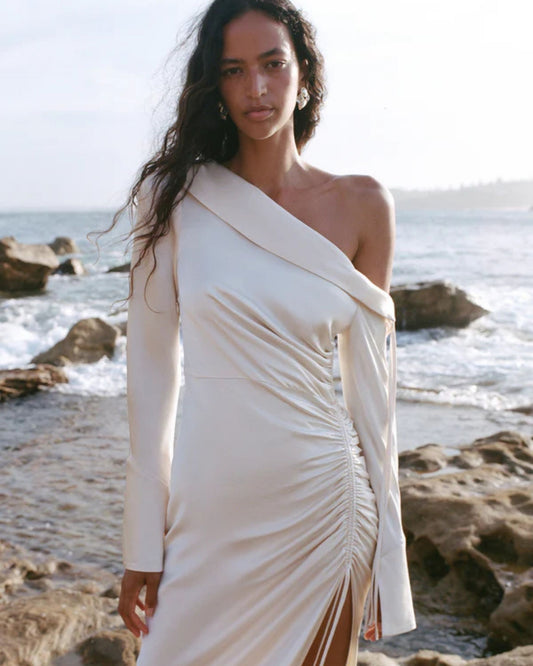 Model wearing a cream satin wedding dress with an asymmetrical neckline, ruched side, cuffed sleeves with ties, and a maxi skirt with a front split. Available at Georgette bridal boutique in Brussels and online at georgettebridal.com.