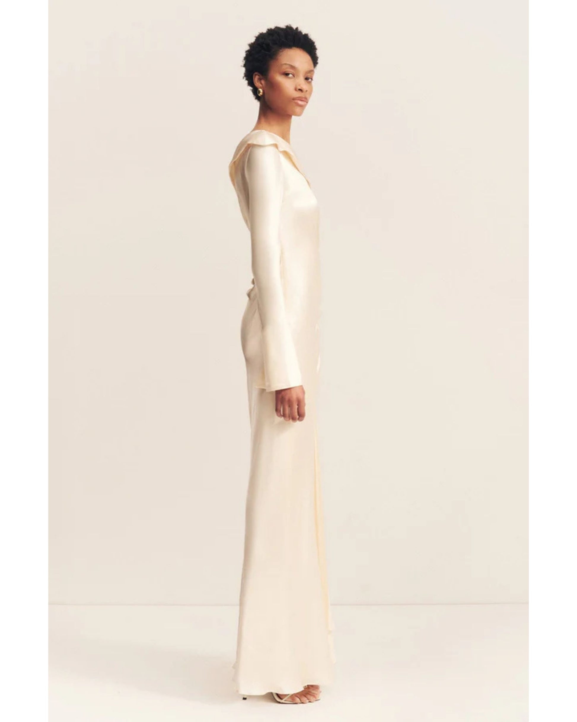 Side view of a cream satin wedding dress with an asymmetrical neckline, ruched side, cuffed sleeves with ties, and a maxi skirt with a front split. Available at Georgette bridal boutique in Brussels and online at georgettebridal.com.