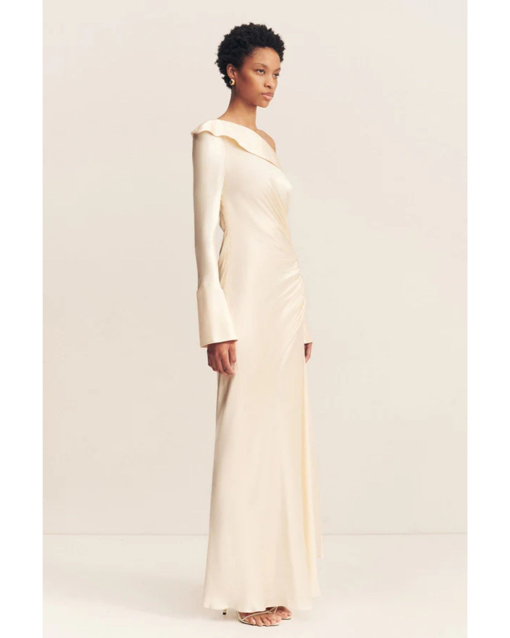 Model wearing a cream satin wedding dress with an asymmetrical neckline, ruched side, cuffed sleeves with ties, and a maxi skirt with a front split. Available at Georgette bridal boutique in Brussels and online at georgettebridal.com.