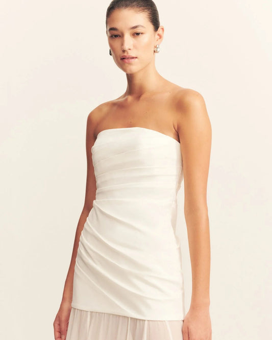 Model wearing a spliced maxi dress with a strapless white stretch cotton blend bodice featuring draped waist tucks, paired with a lightweight, semi-sheer ivory georgette skirt. Available at Georgette bridal boutique in Brussels and online at georgettebridal.com.