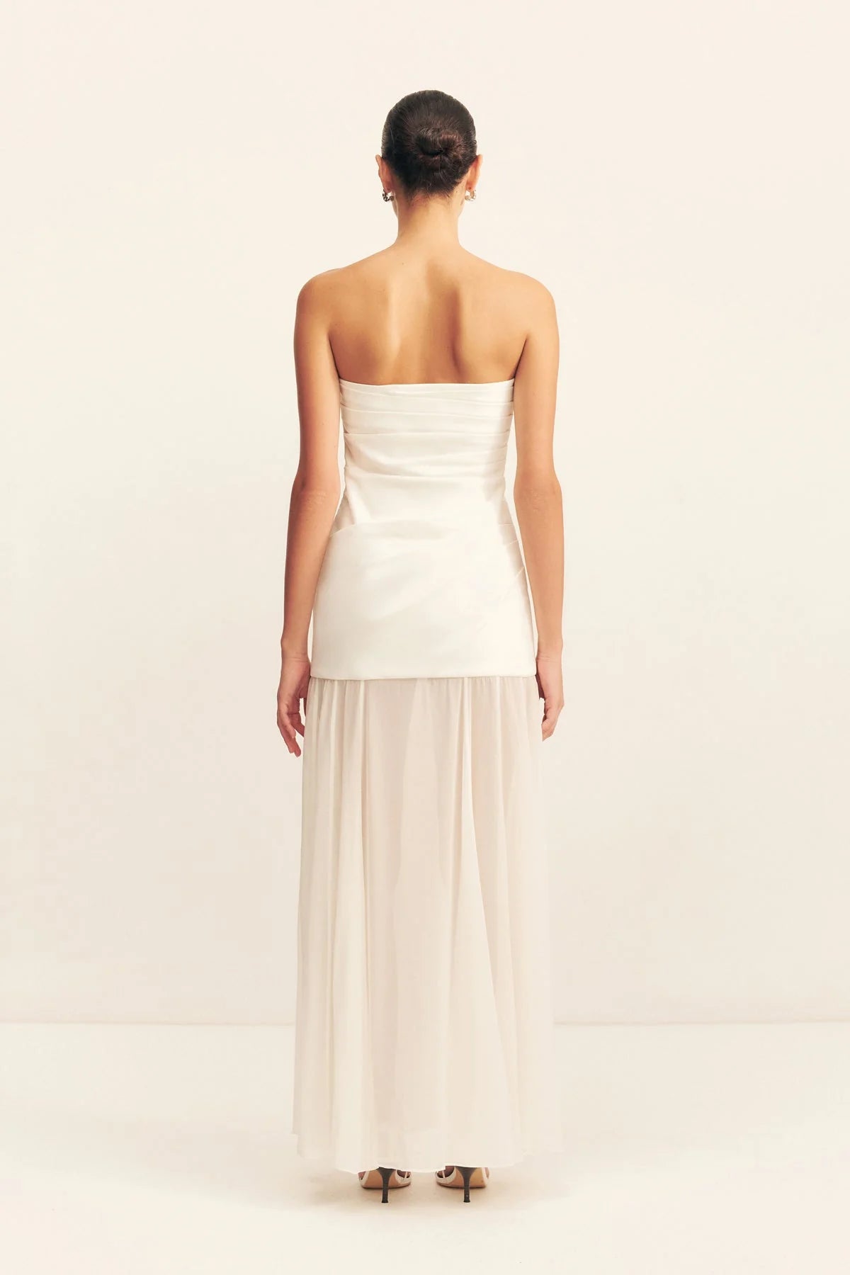Back view, a spliced maxi dress with a strapless white stretch cotton blend bodice featuring draped waist tucks, paired with a lightweight, semi-sheer ivory georgette skirt. Available at Georgette bridal boutique in Brussels and online at georgettebridal.com.