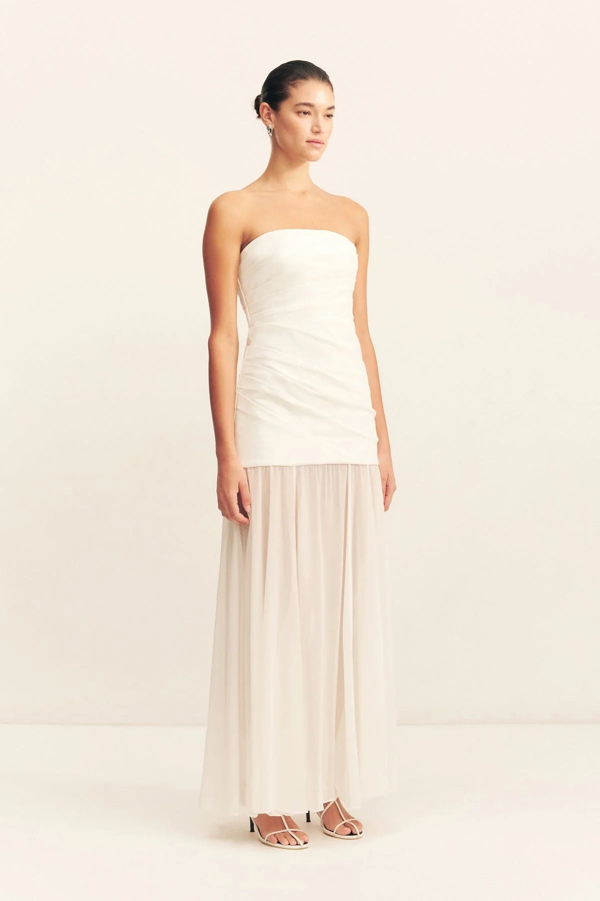 Model wearing a spliced maxi dress with a strapless white stretch cotton blend bodice featuring draped waist tucks, paired with a lightweight, semi-sheer ivory georgette skirt. Available at Georgette bridal boutique in Brussels and online at georgettebridal.com.