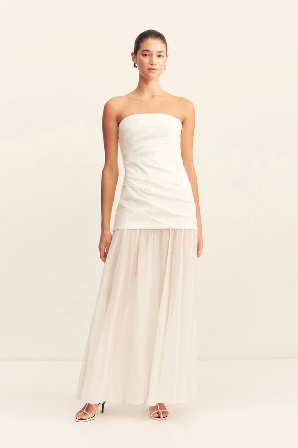 Model wearing a spliced maxi dress with a strapless white stretch cotton blend bodice featuring draped waist tucks, paired with a lightweight, semi-sheer ivory georgette skirt. Available at Georgette bridal boutique in Brussels and online at georgettebridal.com.