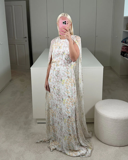 Model wearing the Mina cape dress with a cinched waist and open back, featuring a pastel Buttercups floral print on ivory. The flowing cape can be draped over the shoulders or worn loose, creating an elegant floor-length silhouette. Available at Georgette bridal boutique in Brussels and online at georgettebridal.com.