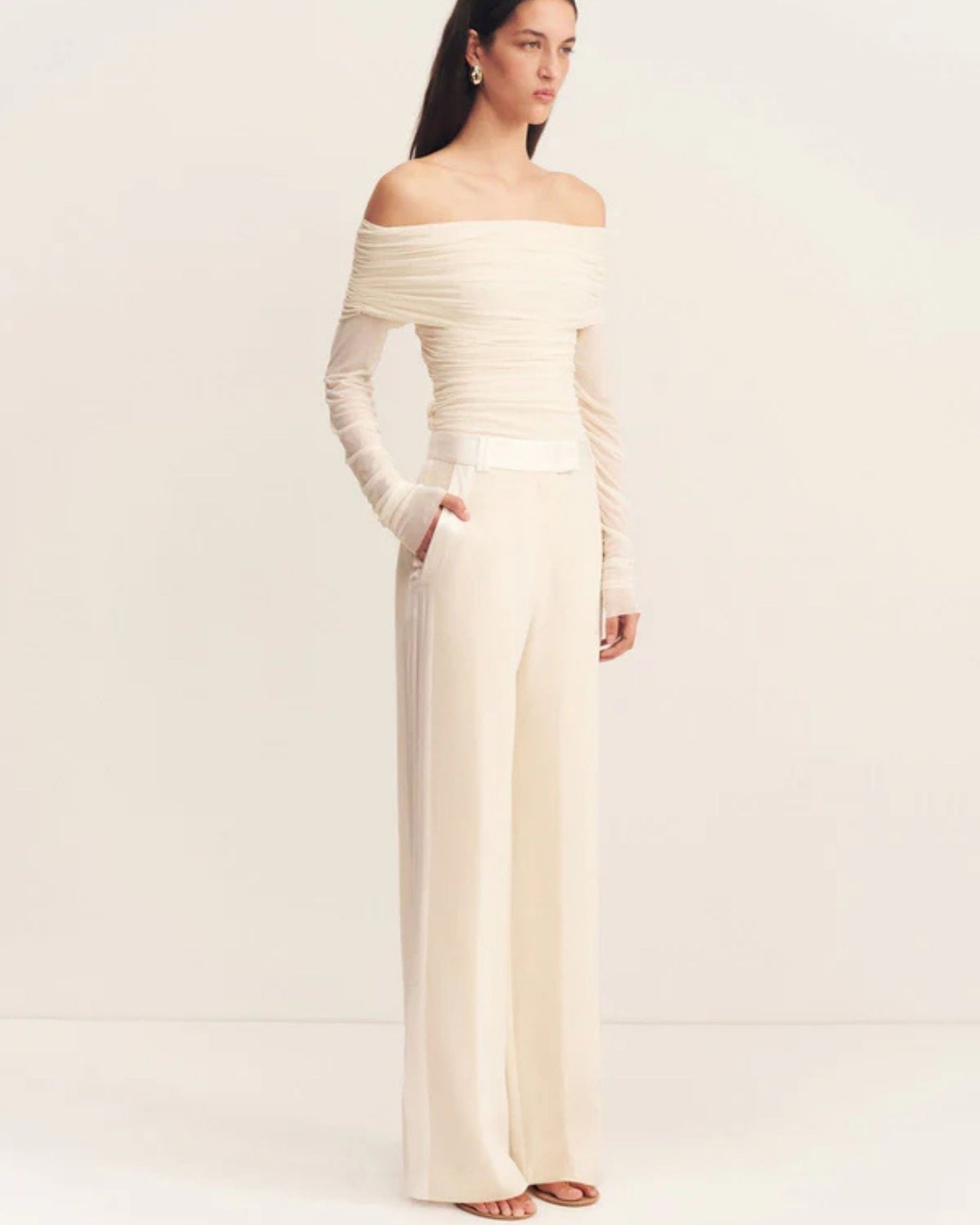 Model wearing the Mira Long Sleeve Off Shoulder Top in ivory - a sheer mesh top with an off-shoulder neckline, long sleeves, and ruched side seams, creating a sculpted silhouette, available at Georgette bridal boutique in Brussels and online at georgettebridal.com.