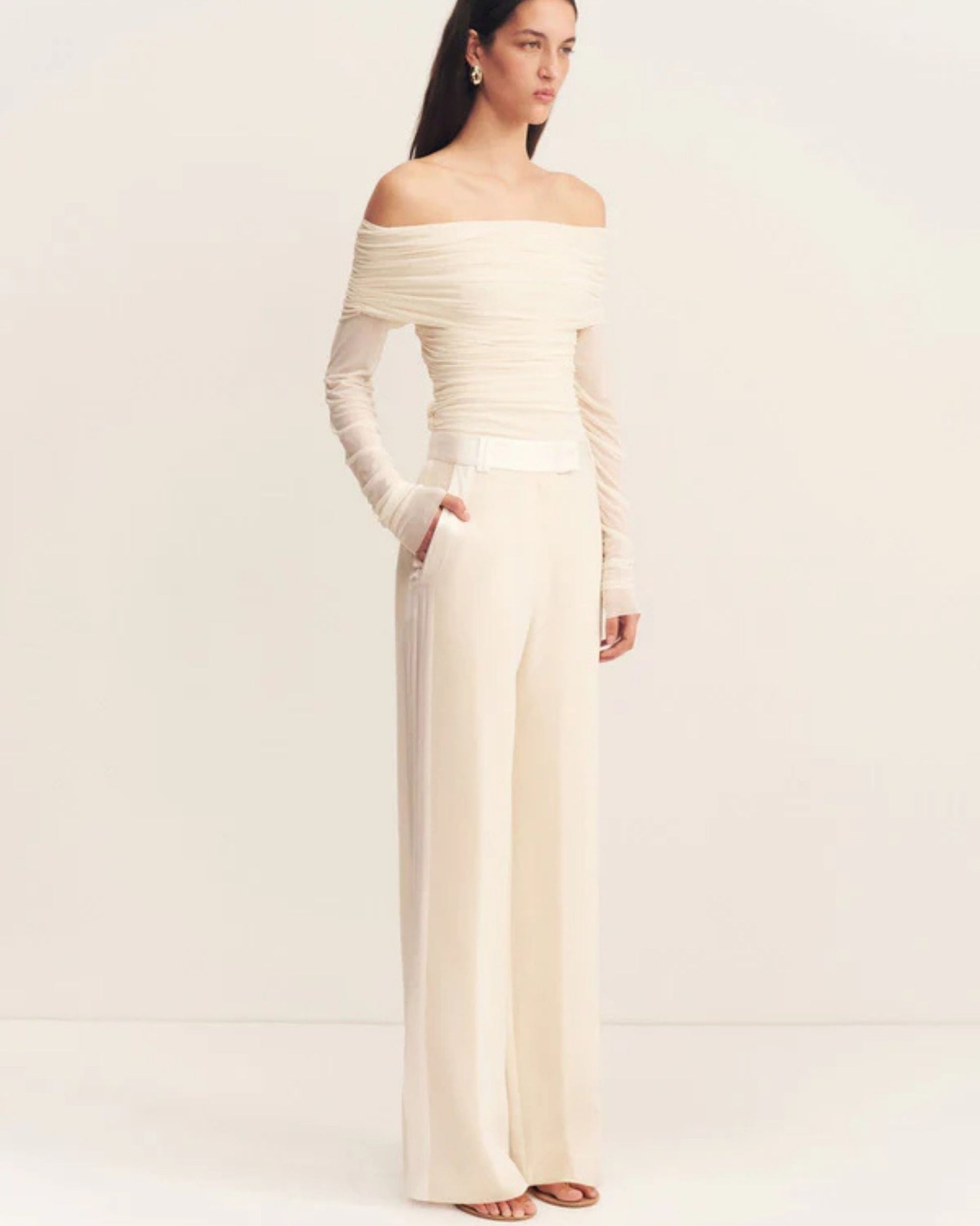 Model wearing the Mira Long Sleeve Off Shoulder Top in ivory - a sheer mesh top with an off-shoulder neckline, long sleeves, and ruched side seams, creating a sculpted silhouette, available at Georgette bridal boutique in Brussels and online at georgettebridal.com.
